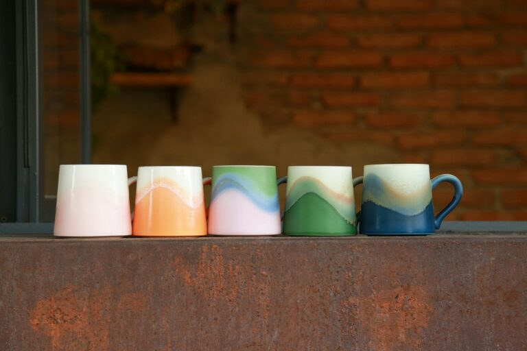Five mugs of the same shape in different glaze colors in a row on a ledge