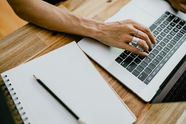 Hand typing on a laptop next to a blank spiral notebook and pencil